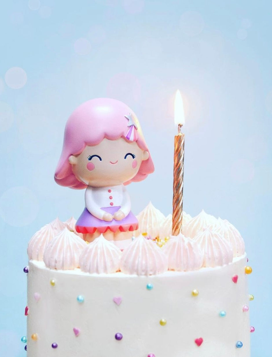 The Birthday Girl Rose Momiji doll cake topper with pink hair and a lit candle sit on a white cake with rainbow sprinkles. — Momiji Doll