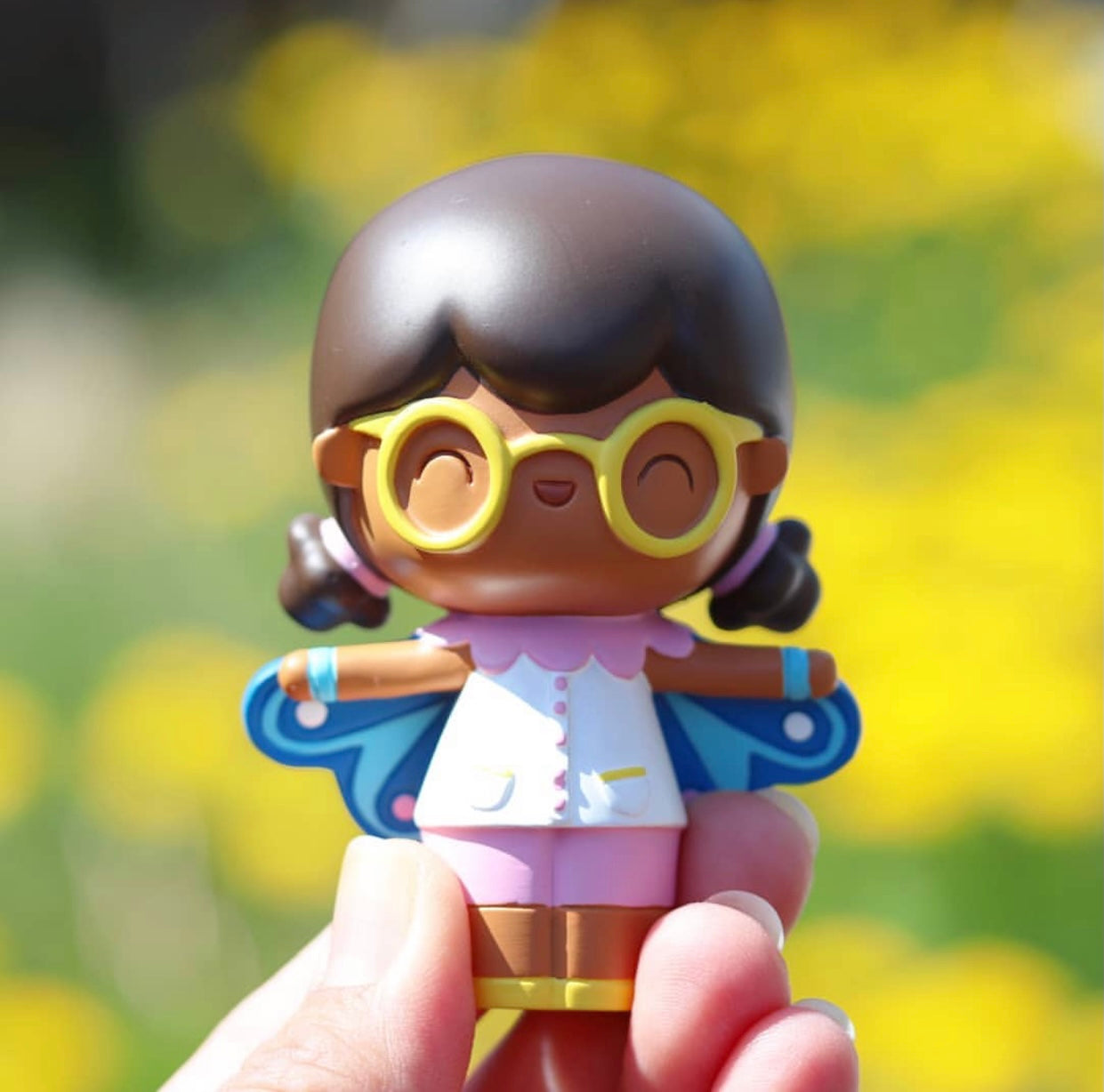 A hand holds Butterfly, a Momiji doll with brown skin, glasses, white outfit, wings, and a smile on a yellow background. — Momiji Doll