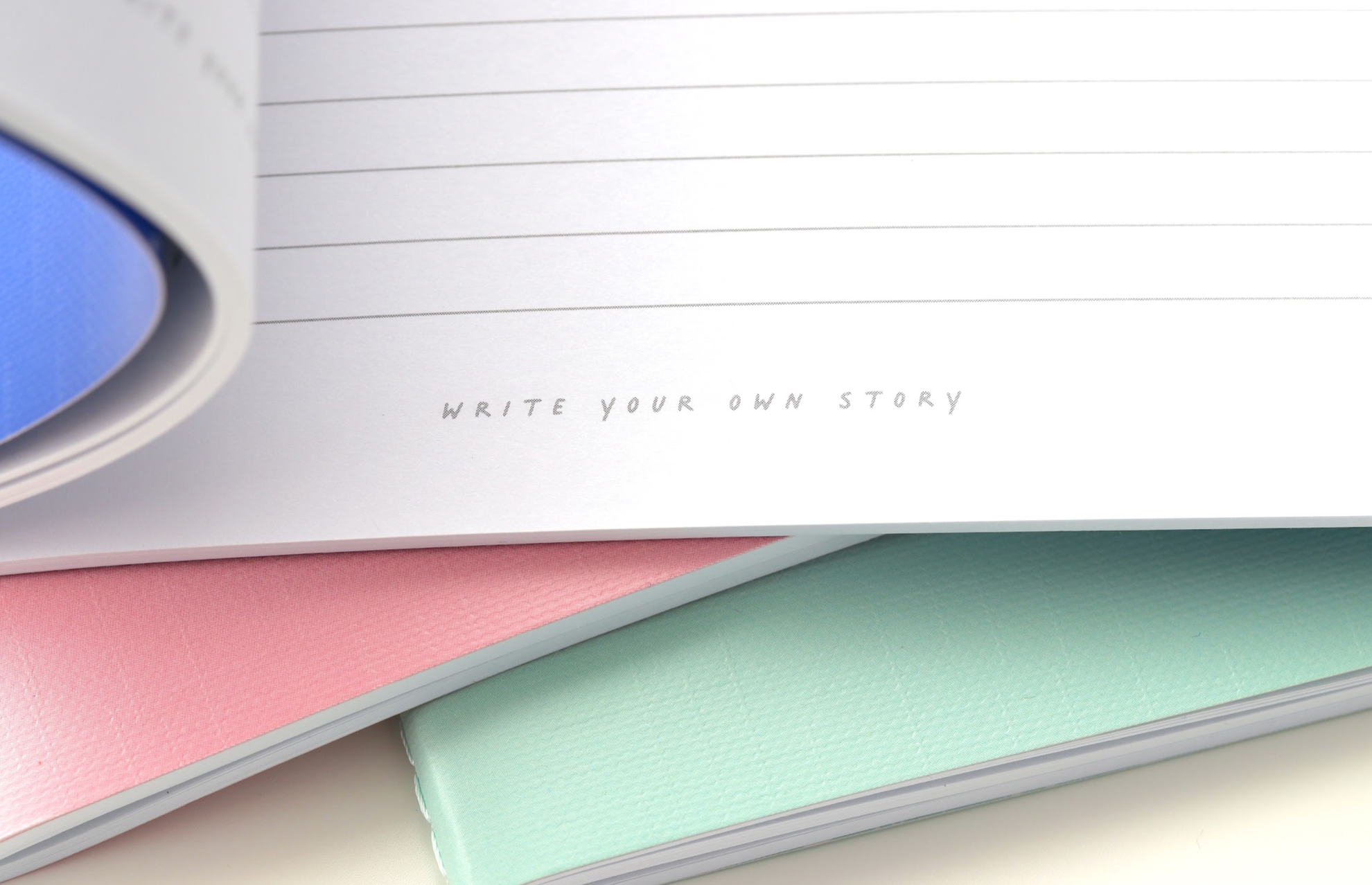 Momiji doll in front of open A5 Going Places Notebook with "WRITE YOUR OWN STORY" on pink, mint, and blue linen covers. — Momiji Doll