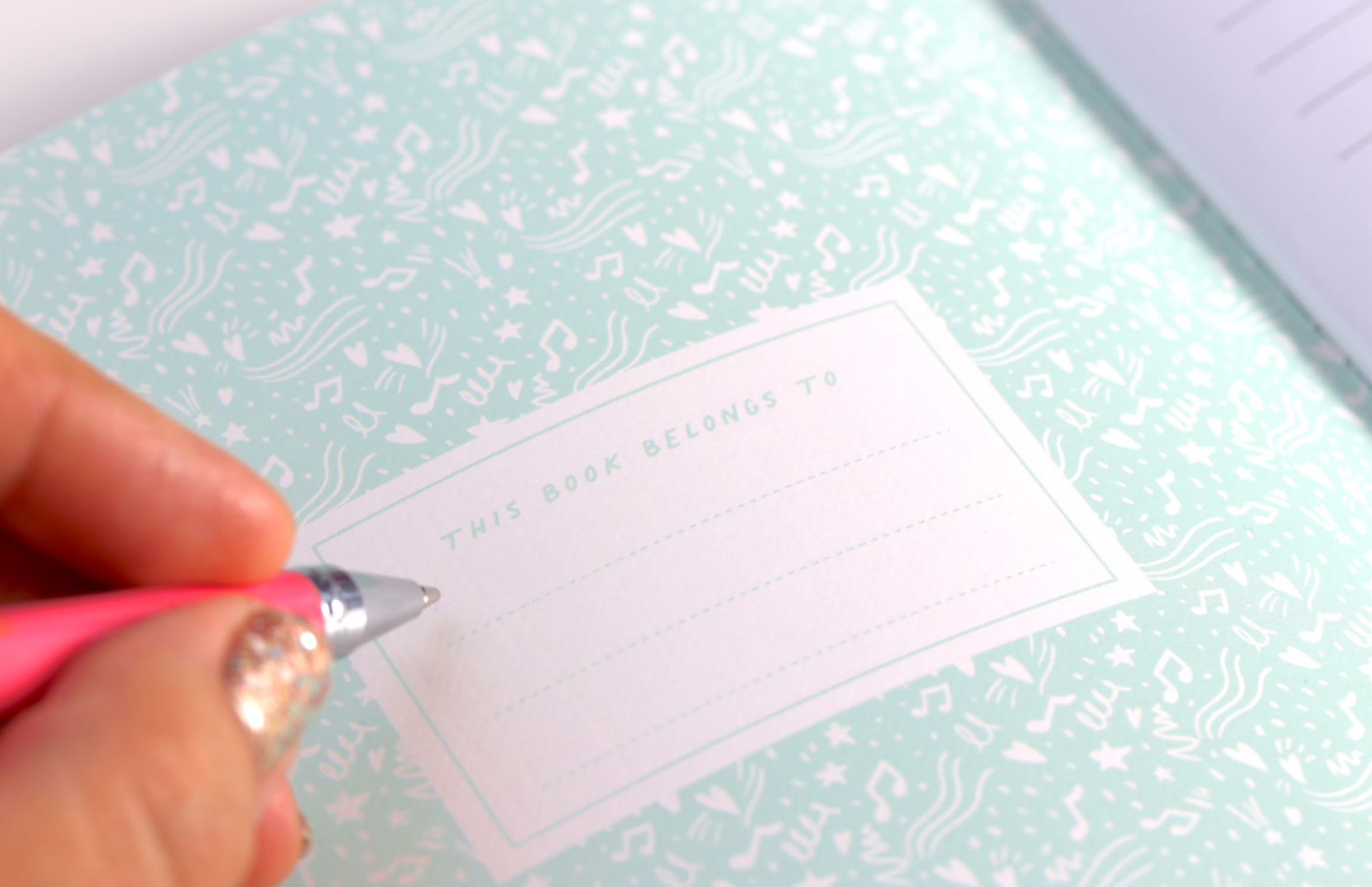 A Momiji doll with the Good Vibes Gang Notebook; a hand holds a pen near the label on its pastel linen A5 cover. — Momiji Doll