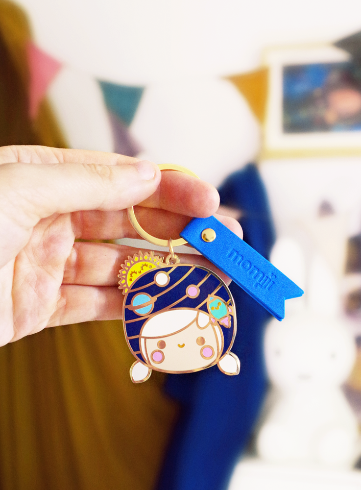 A hand holds the Wonder Momiji doll keychain, a kawaii astronaut charm with gold details and a blue tag in a vibrant room. — Momiji Doll
