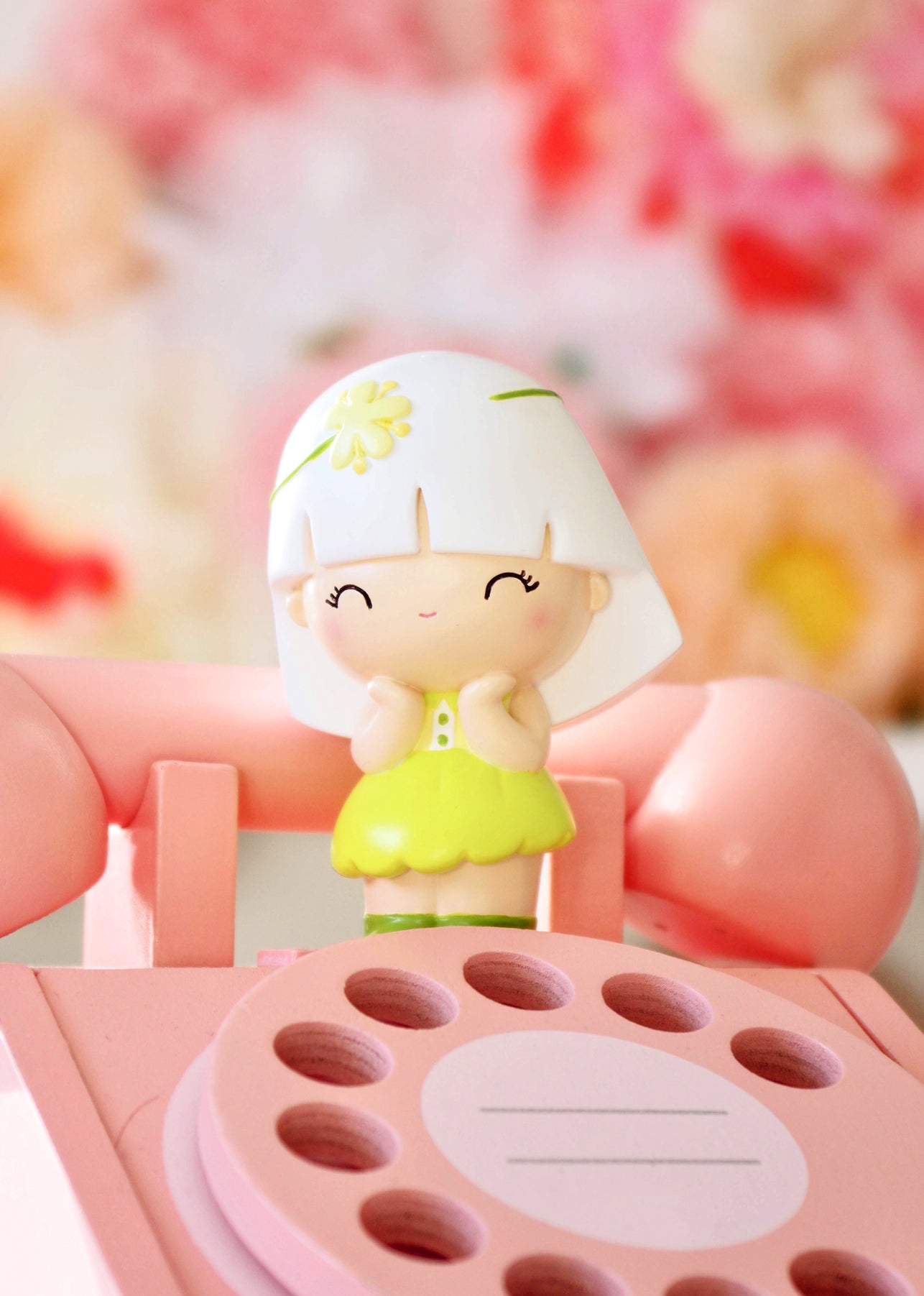 Linden Momiji doll in yellow smiles, standing on a pink phone with floral decor in the background. — Momiji Doll