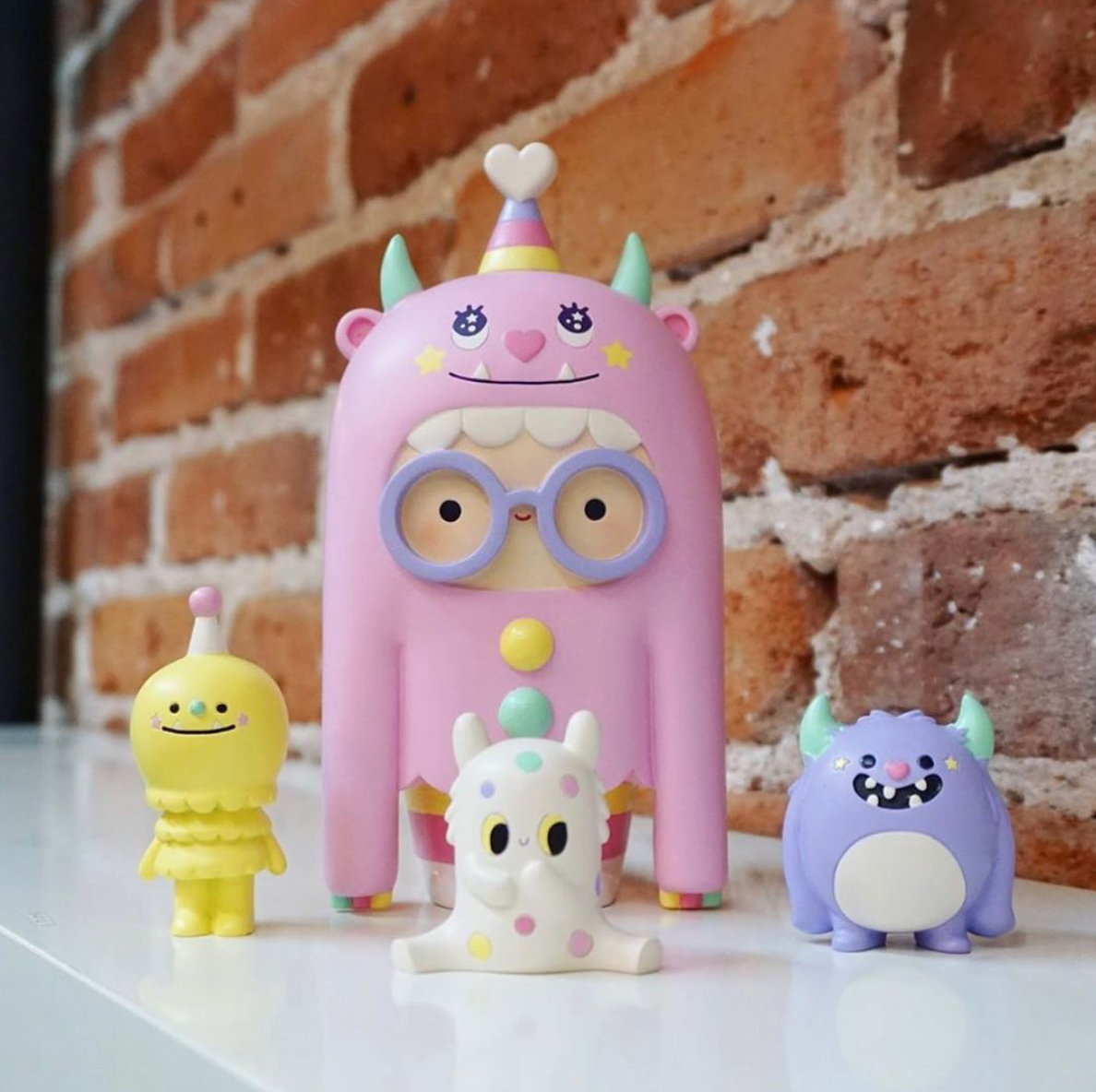 Four colorful Play Big Ideas 2019 Momiji dolls stand on a white surface with a brick wall background. — Momiji Doll