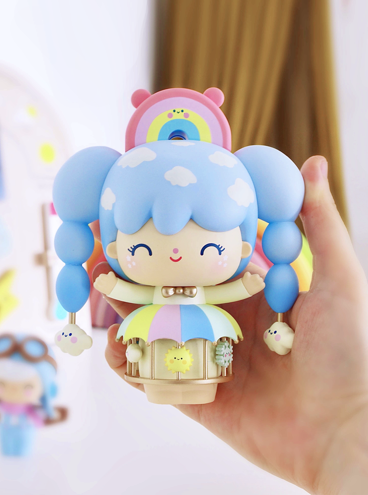 A hand holds a Momiji doll called Sky Dreamer with blue pigtails, rainbow accents, and a pastel cloud dress. — Momiji Doll